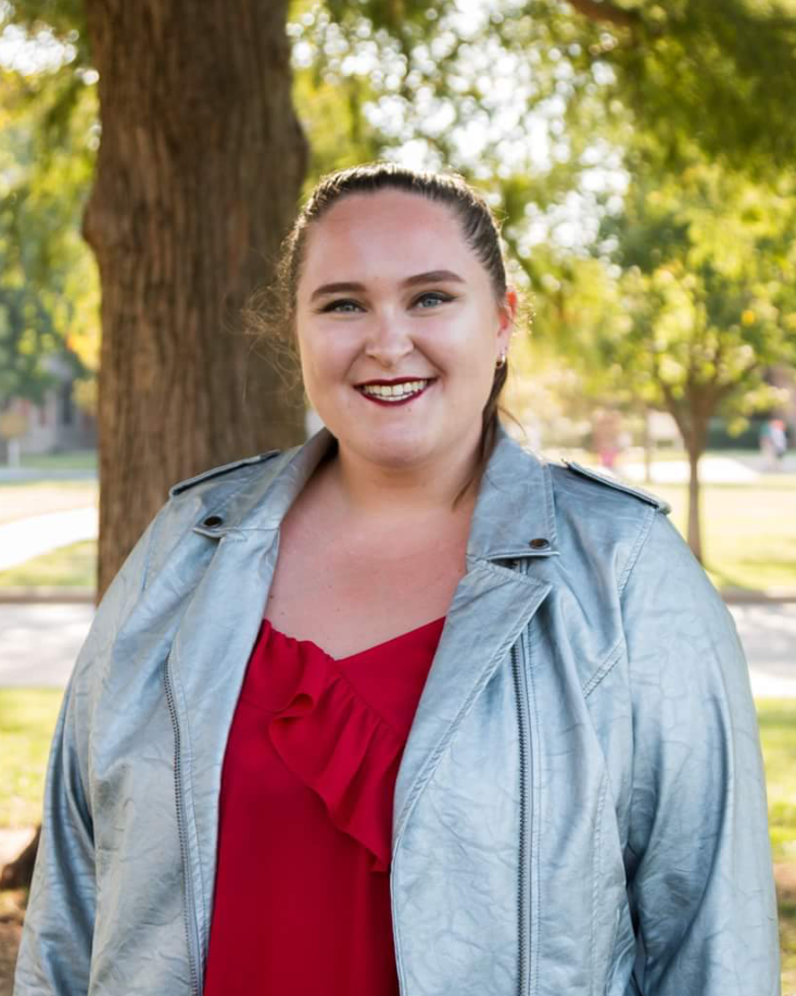 photo of Katy, a white woman with brown hair and red lipstick, outdoors in front of a tree. She is wearing a red shirt and silver jacket.